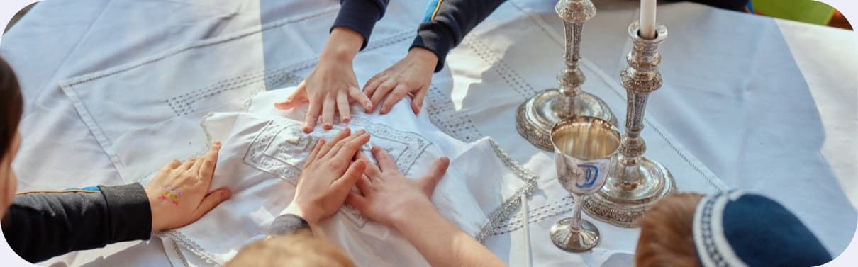 Children placing their hands together on a challah cover beside Shabbat candles